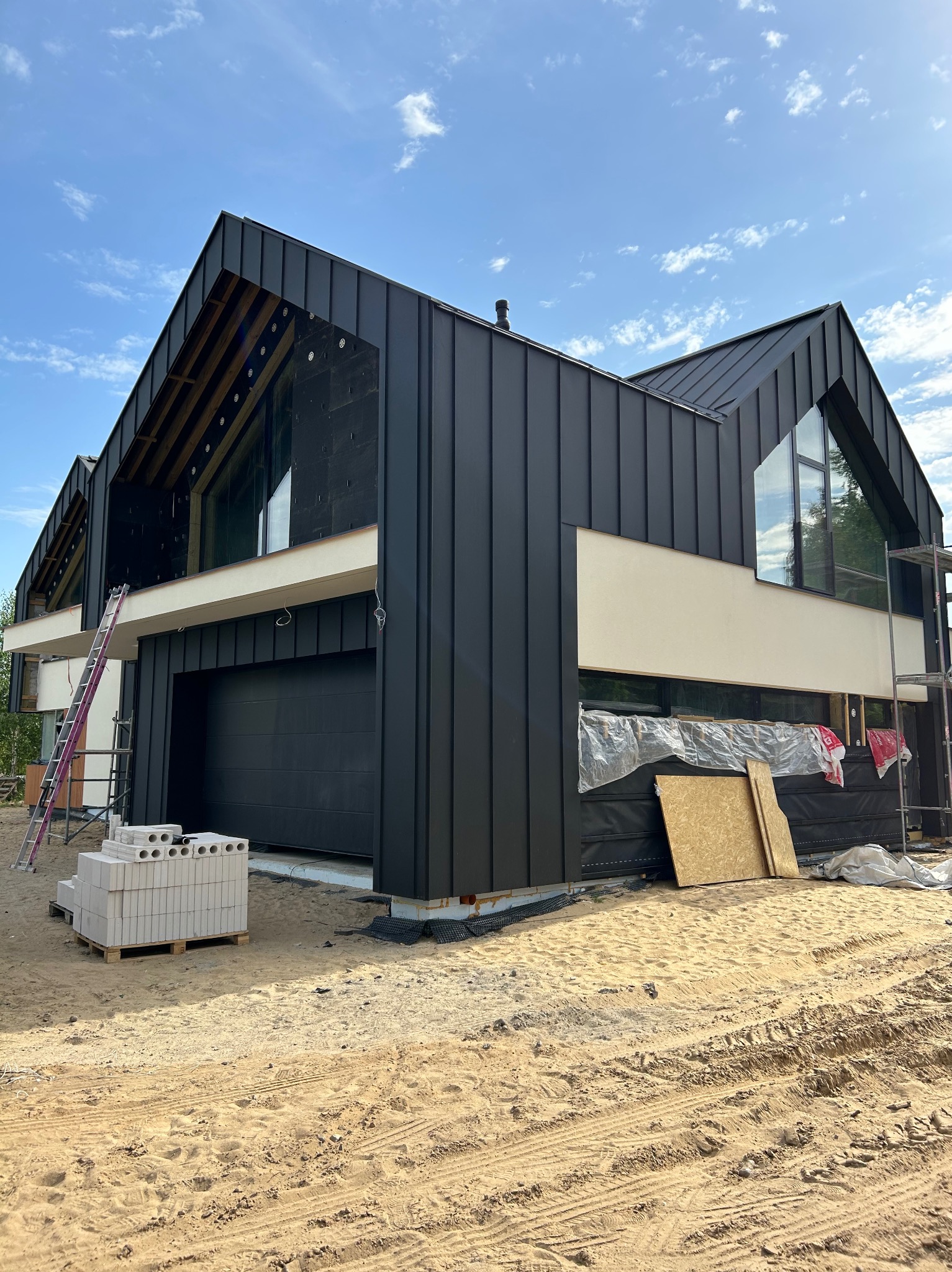 Nowoczesny dom w trakcie budowy z czarną blachą na dachu i ścianach, widoczne rusztowania, paleta z bloczkami i piasek na podwórku.