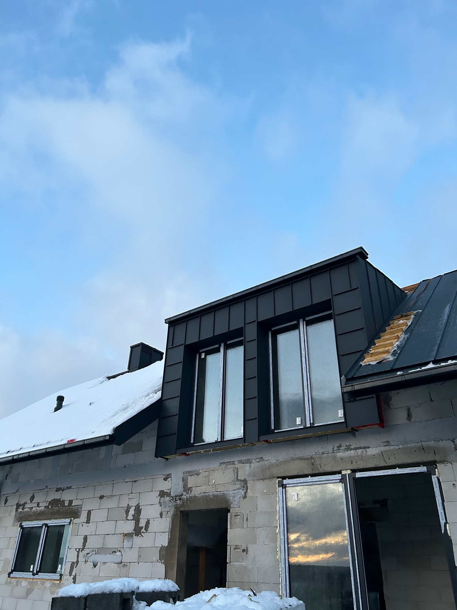 Surowy stan budynku z oknami i dachem pokrytym ciemną blachą, częściowo ośnieżonym. Widoczny fragment dachu z odkrytą konstrukcją drewnianą i kominem.