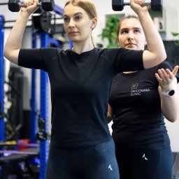 Pracownia Clinic & Formy Warszawa - fizjoterapia - dietetyka - trening medyczny - Kobieta w czarnym stroju sportowym podnosi hantle nad głowę, podczas gdy druga kobieta w czarnej koszulce z logo 'Pracownia Clinic' obserwuje i koryguje jej postawę w siłowni.