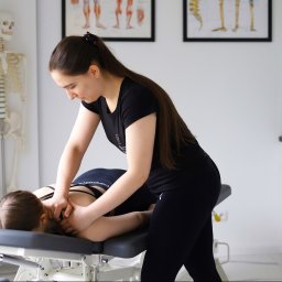 Pracownia Clinic & Formy Warszawa - fizjoterapia - dietetyka - trening medyczny - Kobieta w czarnym stroju masuje szyję pacjentki leżącej na stole rehabilitacyjnym; w tle szkielet i plakaty anatomiczne w gabinecie.