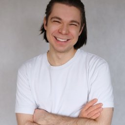 Pracownia Clinic & Formy Warszawa - fizjoterapia - dietetyka - trening medyczny - Uśmiechnięty mężczyzna z ciemnymi włosami, ubrany w biały t-shirt, stoi z założonymi rękami na tle szarej ściany.