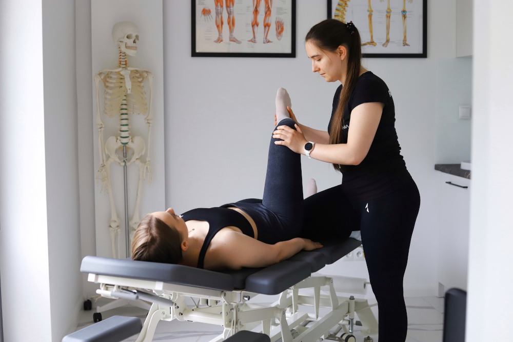 Fizjoterapeutka w czarnym stroju rozciąga nogę pacjentki leżącej na profesjonalnym stole rehabilitacyjnym; w tle model szkieletu i plansze anatomiczne.