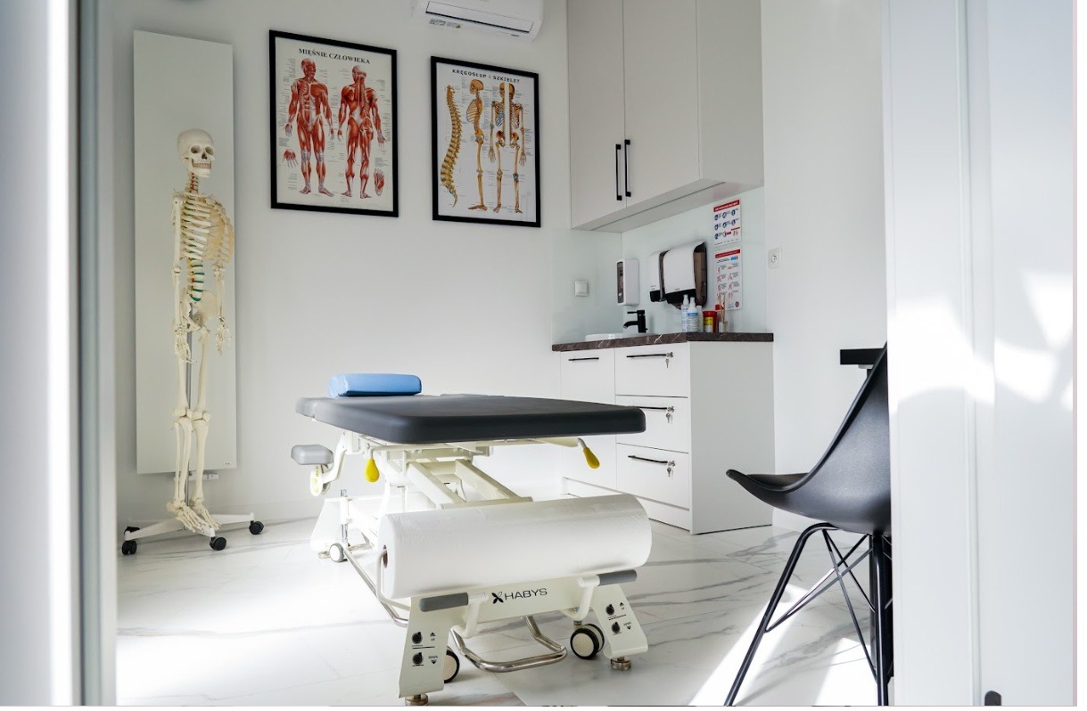 Jasne wnętrze gabinetu z regulowanym stołem do masażu, modelem szkieletu i plakatami anatomicznymi na ścianie.