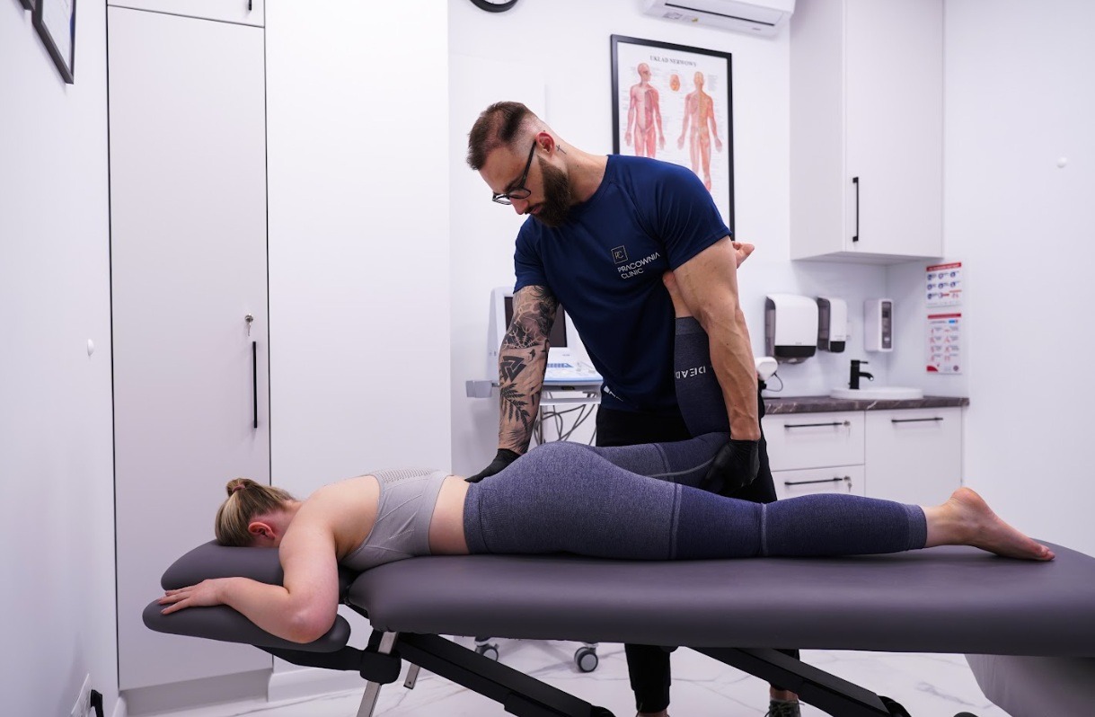 Kobieta leżąca na stole rehabilitacyjnym podczas terapii manualnej wykonywanej przez mężczyznę z tatuażami na ramieniu, w tle plakat z układem nerwowym człowieka i białe szafki.