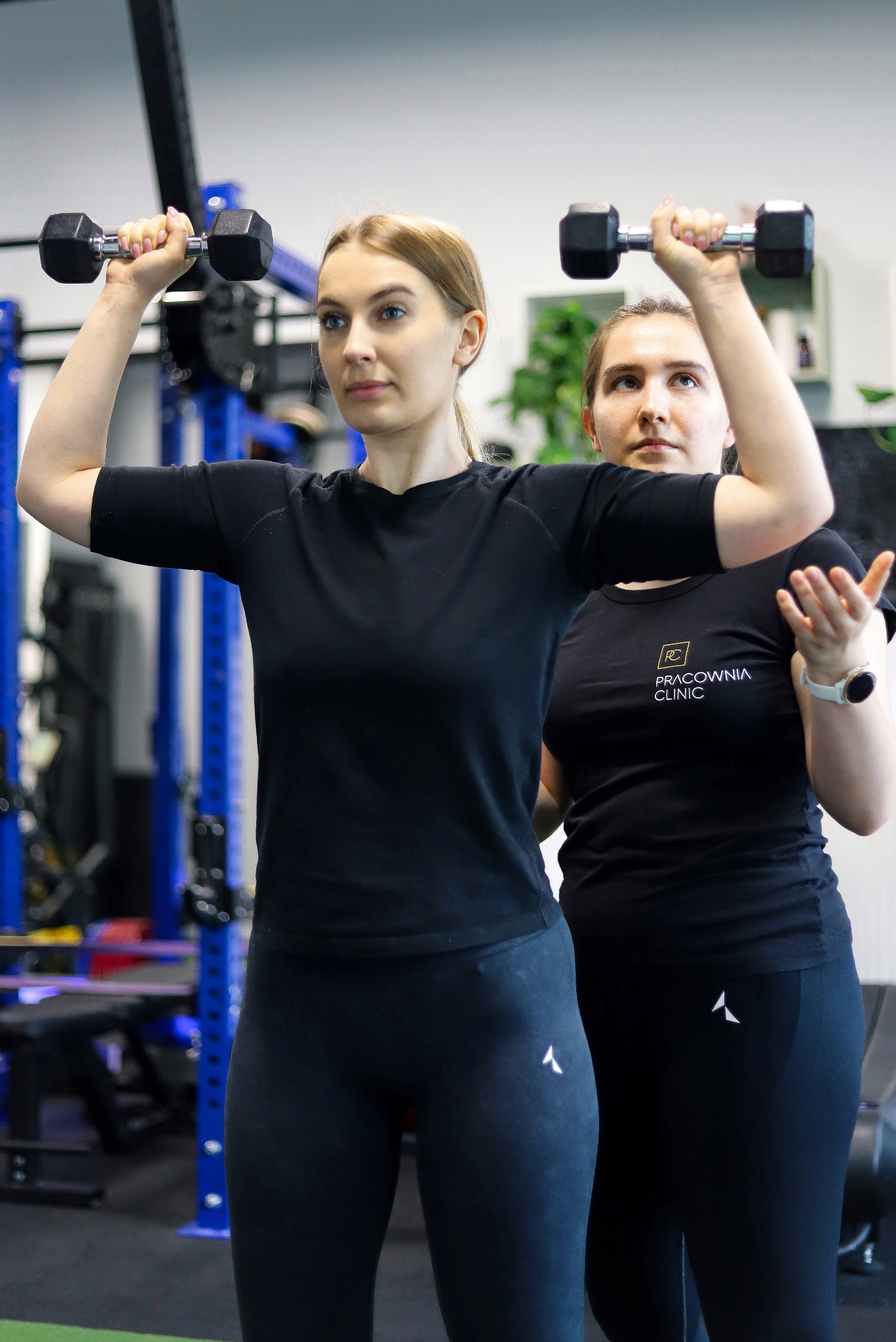 Kobieta w czarnym stroju sportowym podnosi hantle nad głowę, podczas gdy druga kobieta w czarnej koszulce z logo 'Pracownia Clinic' obserwuje i koryguje jej postawę w siłowni.
