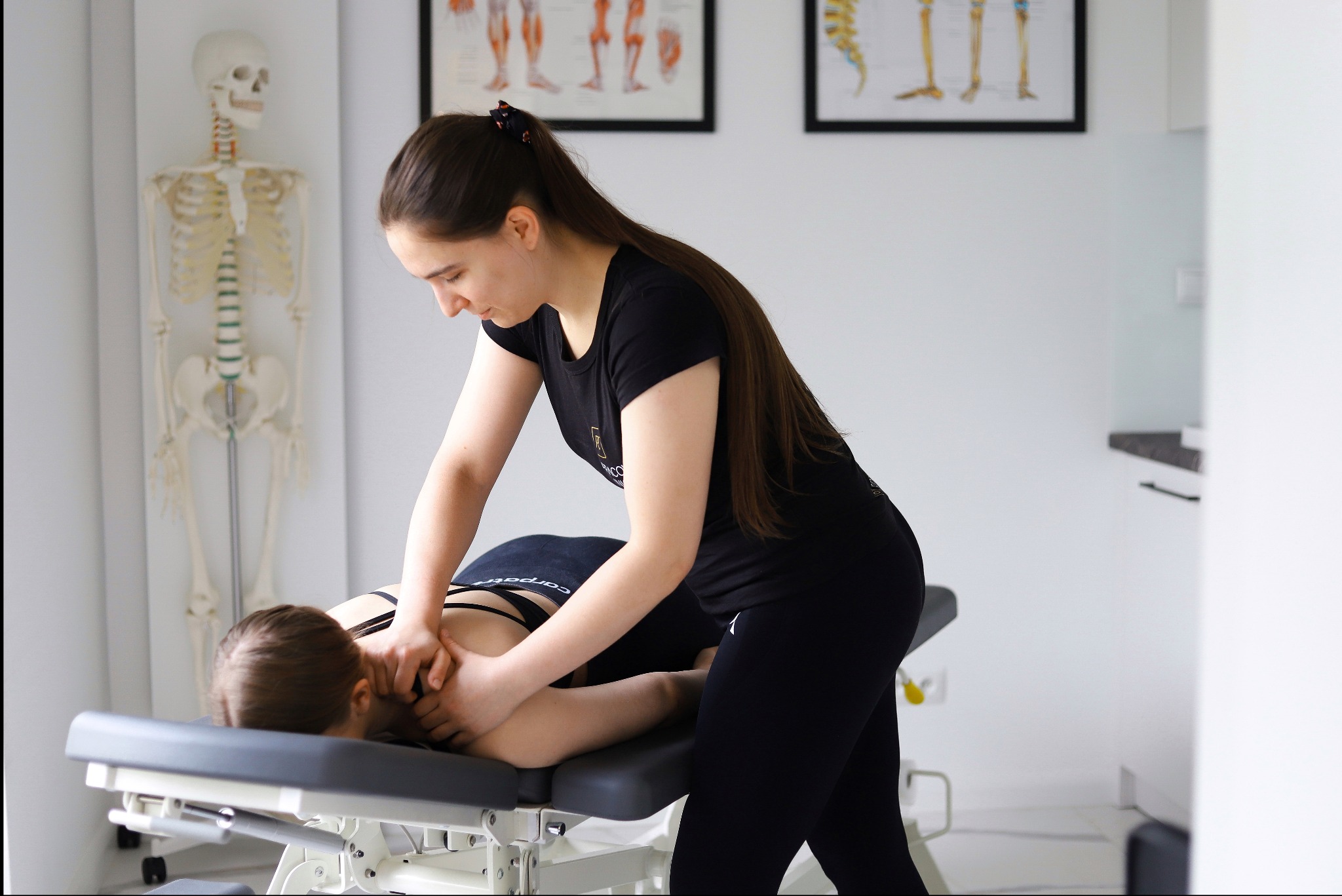 Kobieta w czarnym stroju masuje szyję pacjentki leżącej na stole rehabilitacyjnym; w tle szkielet i plakaty anatomiczne w gabinecie.