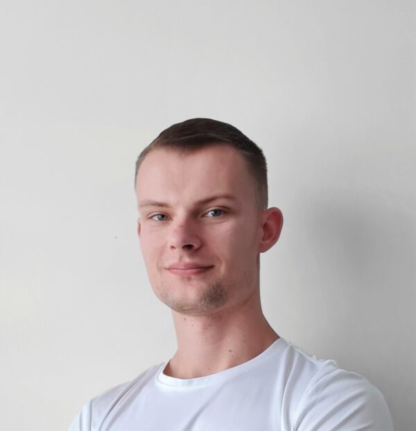 Portret młodego mężczyzny w białej koszulce, z lekkim uśmiechem i małym zadrapaniem na nosie, na jasnym, neutralnym tle.