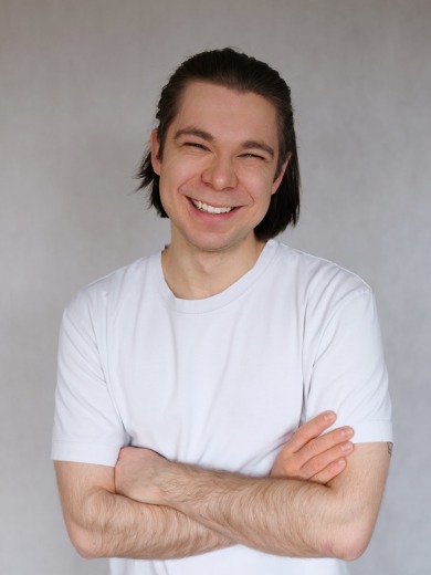 Uśmiechnięty mężczyzna z ciemnymi włosami, ubrany w biały t-shirt, stoi z założonymi rękami na tle szarej ściany.