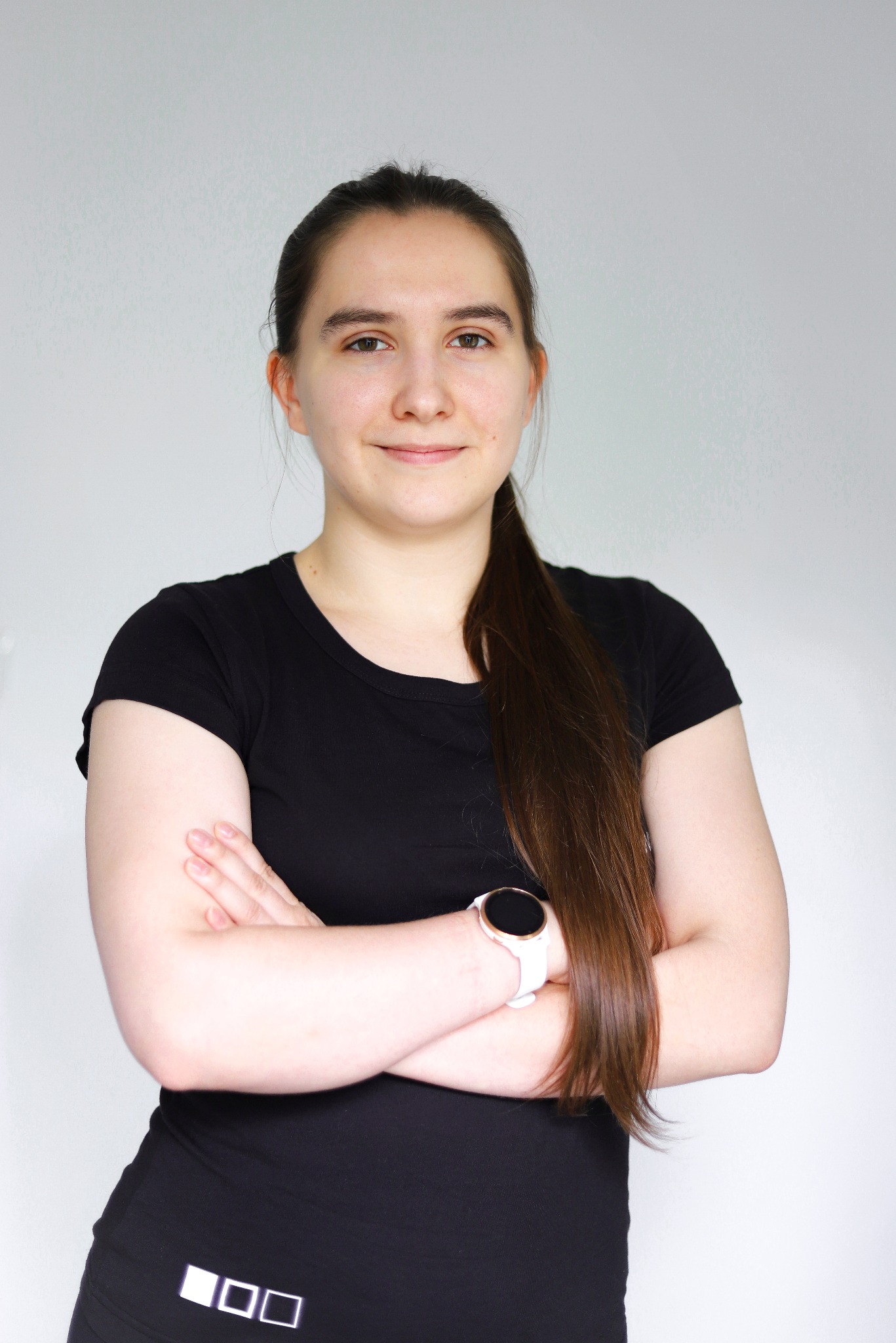 Portret uśmiechniętej kobiety w czarnej koszulce sportowej z białym zegarkiem na ręce, stojącej na jasnym tle z założonymi rękami.