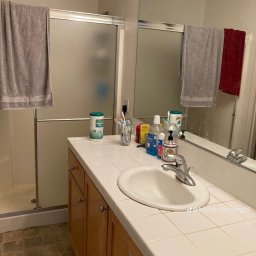 BioCleanMaster - Umywalka w łazience z białym blatem wyłożonym kafelkami, nad nią lustro z odbiciem ręczników i kabiny prysznicowej, obok widoczne środki czystości i kosmetyki.