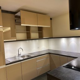 BioCleanMaster - Beżowa, nowoczesna kuchnia z szarym blatem i podświetleniem LED pod szafkami. Widoczny zlew, bateria i płyta indukcyjna. Uwagę przyciąga otwierana szafka górna.