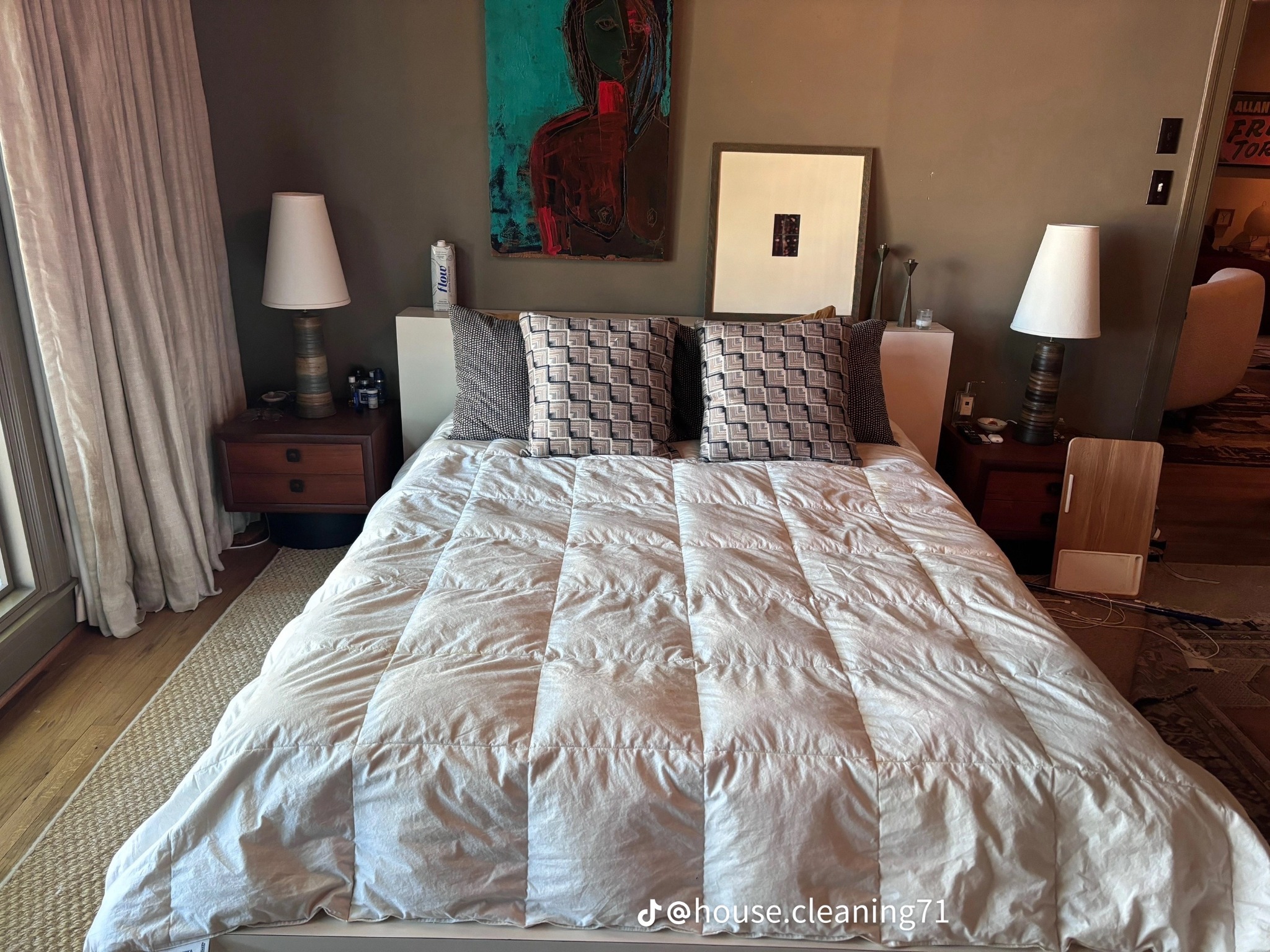 Sypialnia z dużym łóżkiem z białą kołdrą, wzorzyste poduszki, stoliki nocne z lampkami, abstrakcyjny obraz na ścianie, drewniana podłoga i dywan.