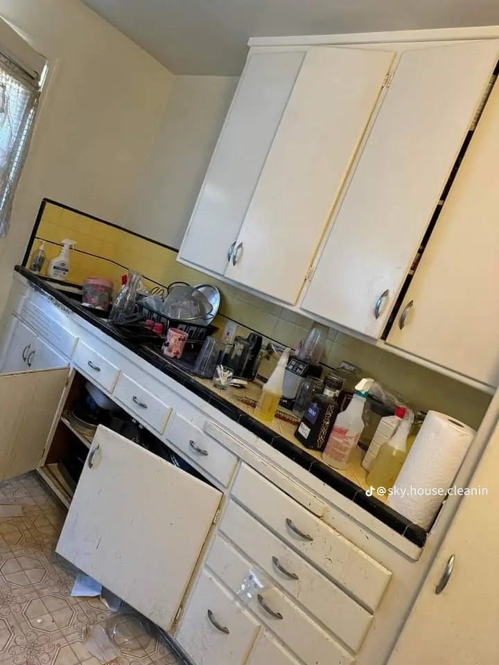 Zaniedbana kuchnia z otwartymi szafkami, blat zastawiony przedmiotami, widoczny wzorzysty linoleum na podłodze i okno z firanką.