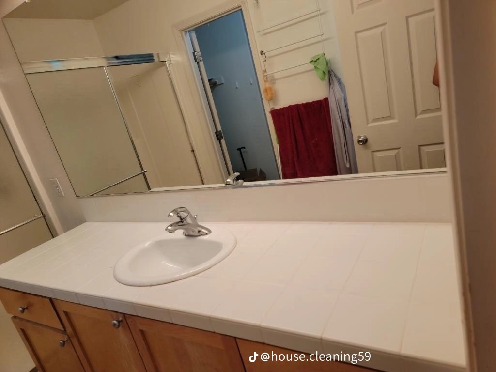 Umywalka z białym blatem w łazience, widoczne odbicie w lustrze z suszącymi się ręcznikami i otwartymi drzwiami prysznica.