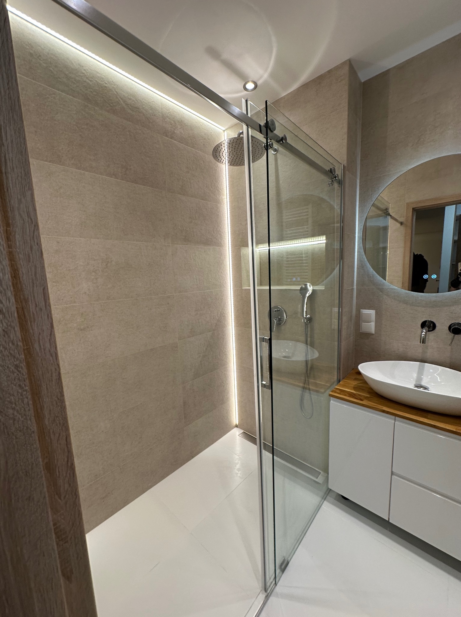 Nowoczesna łazienka z kabiną prysznicową typu walk-in, oświetleniem LED i okrągłym lustrem z podświetleniem. Umywalka nablatowa na szafce z drewnianym blatem.