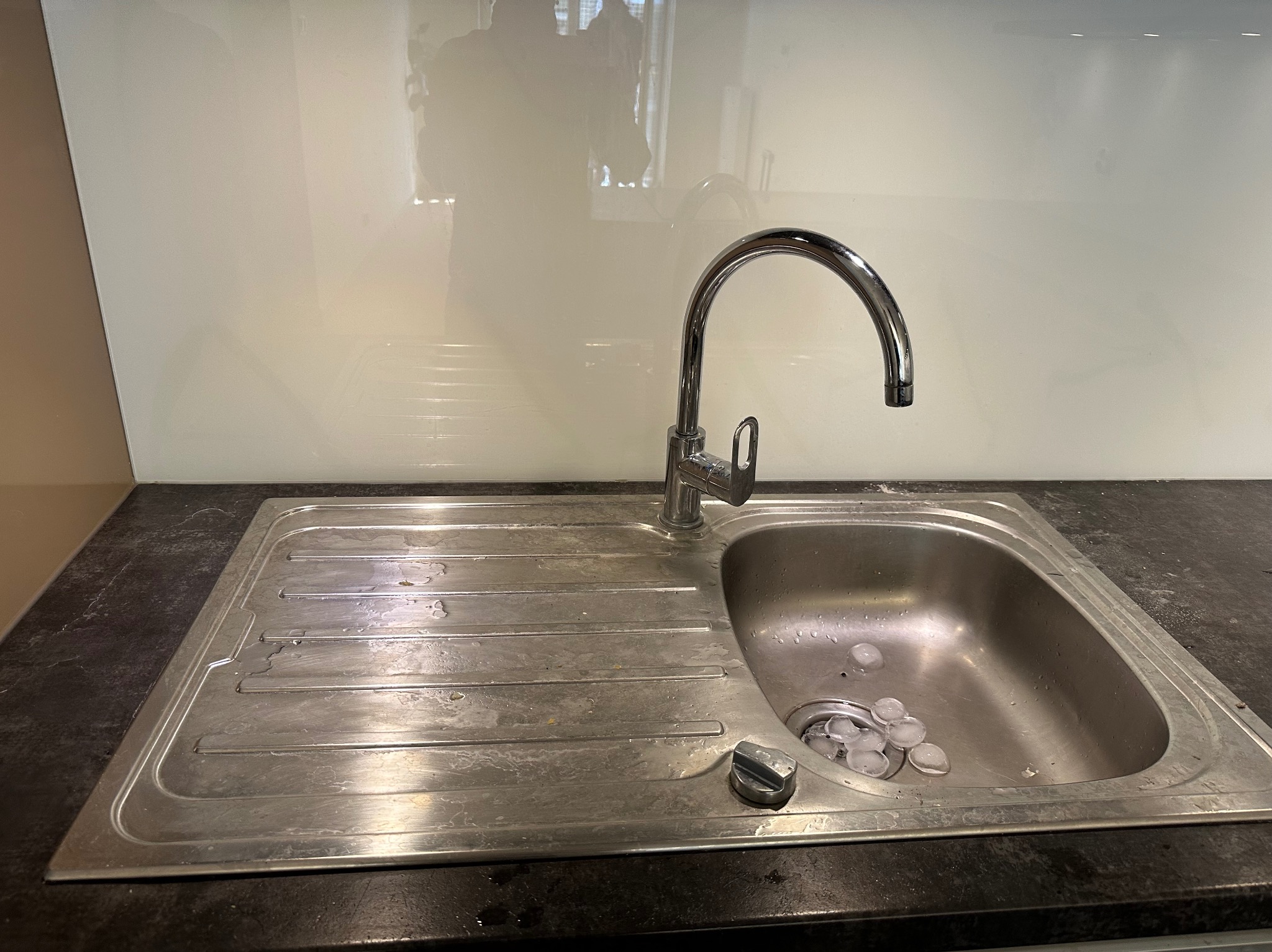 Zlew kuchenny ze stali nierdzewnej przed czyszczeniem, widoczne zabrudzenia i osad. W odpływie leżą kostki lodu. Bateria chromowana, tło stanowi jasny panel ścienny.