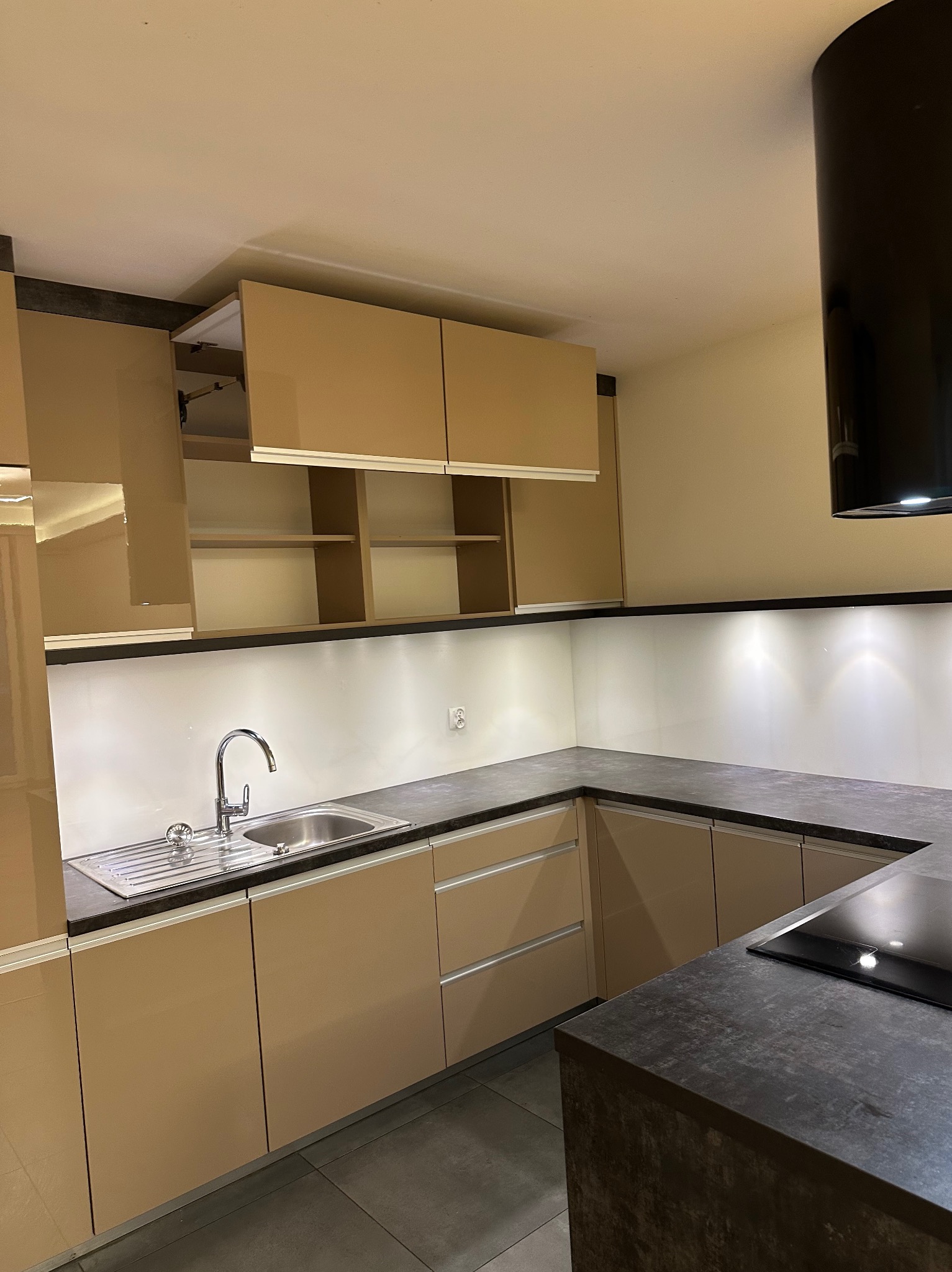 Nowoczesna, beżowa kuchnia z ciemnym blatem i oświetleniem LED. Widok na zlew, baterię i płytę indukcyjną. Minimalistyczny design z otwartą szafką.