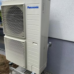 Doko Sp. Z o.o. - Zewnętrzna jednostka klimatyzacji Panasonic zamontowana na metalowej konstrukcji Elektromatic przy ścianie budynku w Brodnicy, z widocznym przewodem odprowadzającym skropliny.