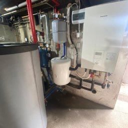 Doko Sp. Z o.o. - Biały, wiszący piec grzewczy Panasonic z podłączonymi rurami i zbiornikami w kotłowni. Widoczne liczne rury, zawory i zbiorniki wyrównawcze. Niska jakość wykończenia pomieszczenia.