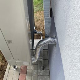 Doko Sp. Z o.o. - Zewnętrzna jednostka klimatyzacji zamontowana na elewacji budynku, z widocznym izolowanym przewodem przechodzącym przez ścianę, ułożona kostka brukowa.