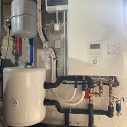 Doko Sp. Z o.o. - Zainstalowana biała pompa ciepła Panasonic Aquarea z widocznymi rurami w izolacji termicznej i zbiornikiem buforowym na tle surowej ściany.