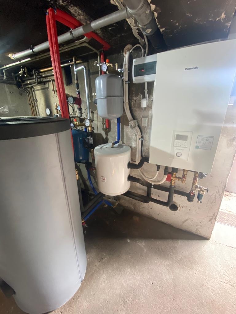 Biały, wiszący piec grzewczy Panasonic z podłączonymi rurami i zbiornikami w kotłowni. Widoczne liczne rury, zawory i zbiorniki wyrównawcze. Niska jakość wykończenia pomieszczenia.
