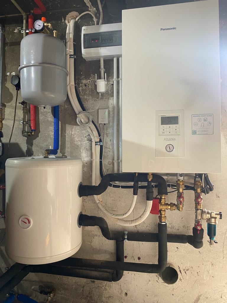 Zainstalowana biała pompa ciepła Panasonic Aquarea z widocznymi rurami w izolacji termicznej i zbiornikiem buforowym na tle surowej ściany.