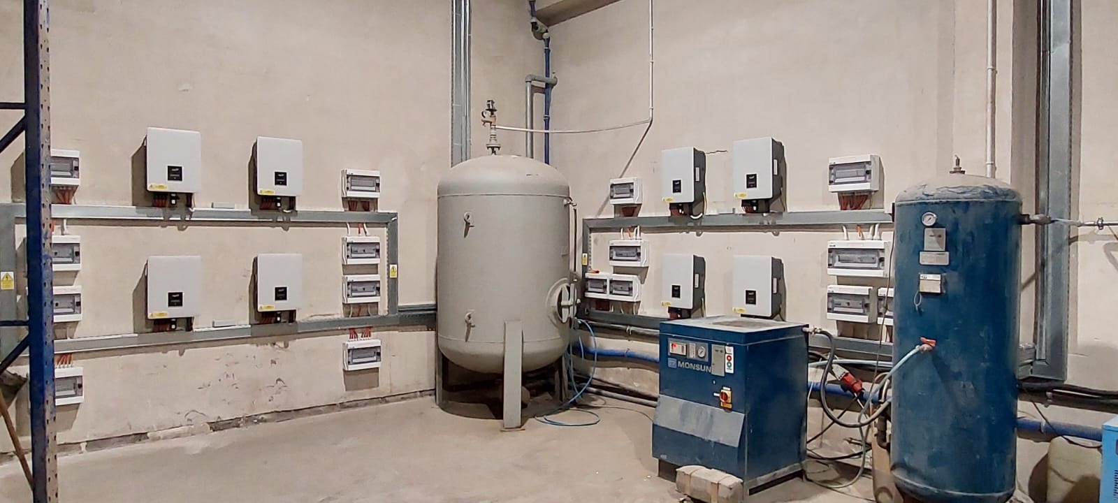 Rząd falowników fotowoltaicznych i skrzynek elektrycznych zamontowanych na ścianie w pomieszczeniu technicznym, obok zbiorników ciśnieniowych i sprężarki powietrza marki Monsun.