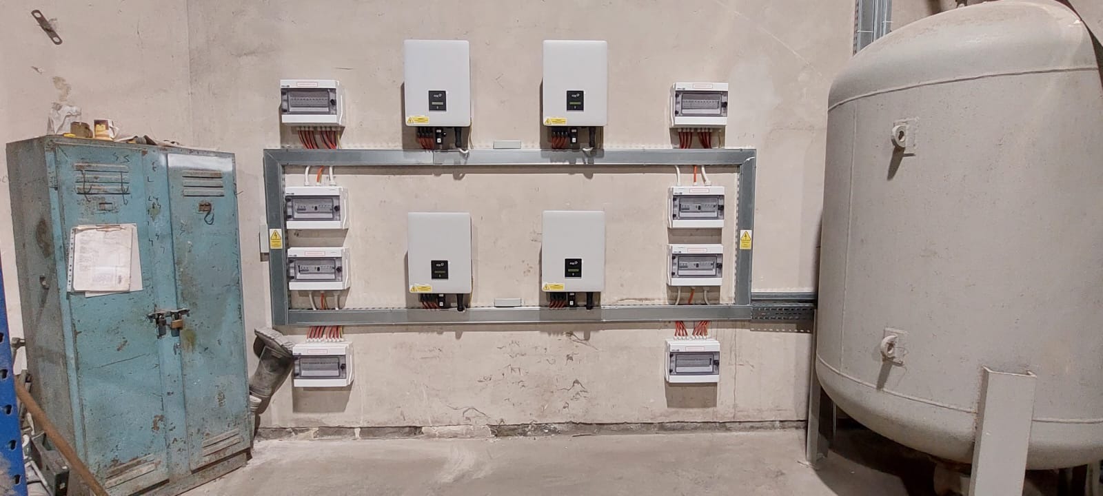 Ściana z zamontowanymi inwerterami i rozdzielnicami połączonymi metalowymi szynami, widoczny również stary, metalowy schowek i zbiornik, surowy stan wykończenia wnętrza.