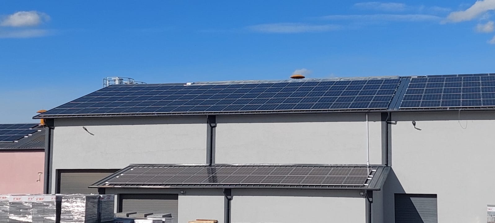 Panele słoneczne na dachu budynku przemysłowego z widocznym kominem wentylacyjnym i paletami materiałów budowlanych w dolnej części kadru.