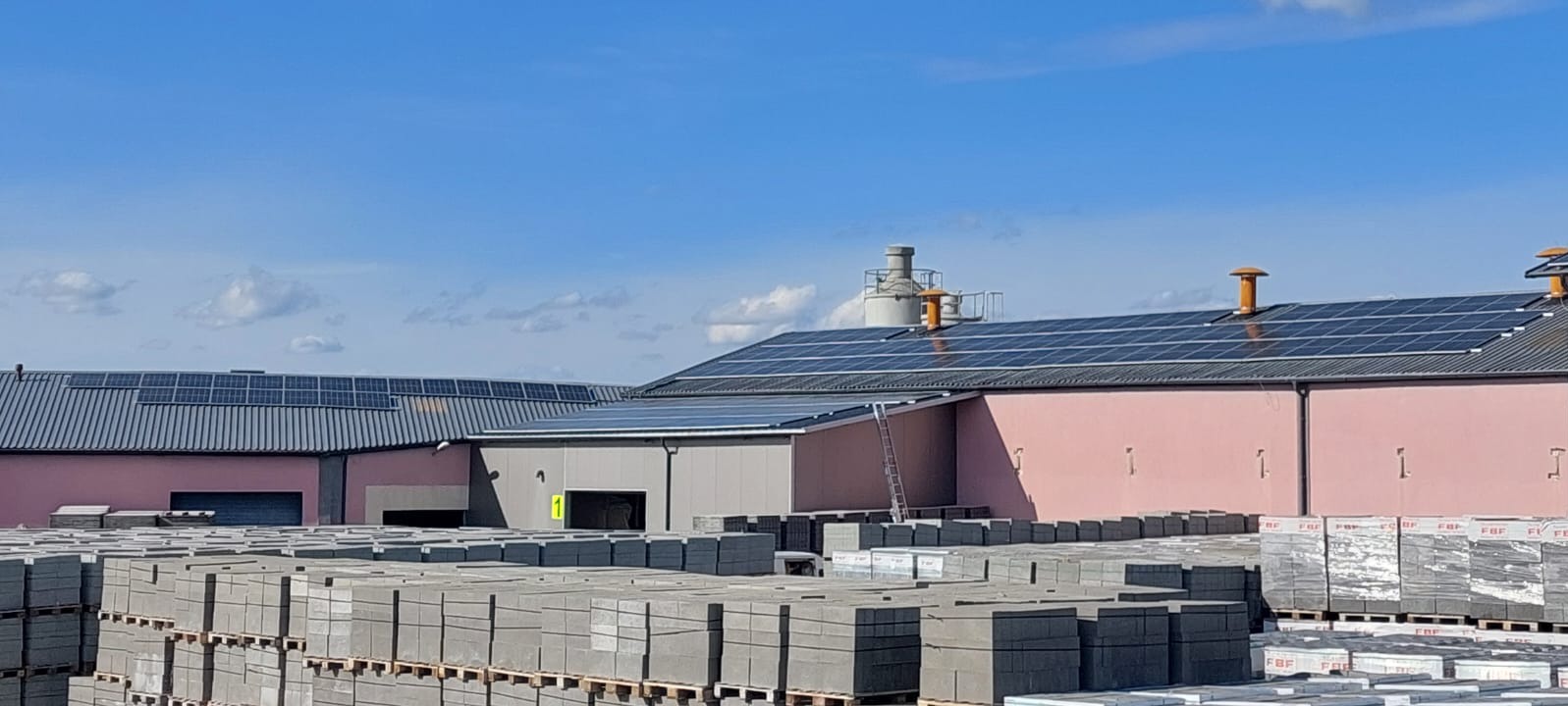 Budynki przemysłowe z panelami słonecznymi na dachu, na pierwszym planie ułożone palety z kostką brukową.