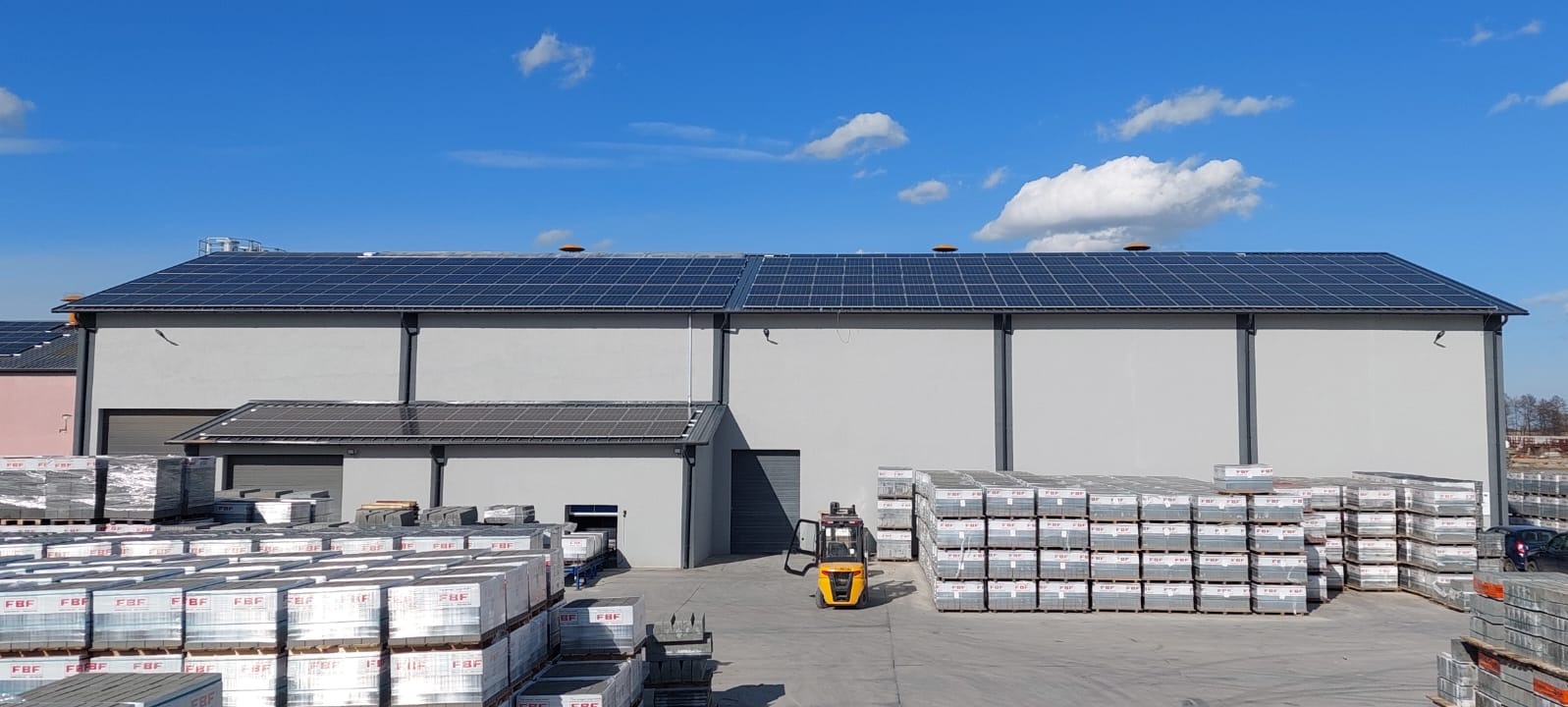 Hala magazynowa z panelami słonecznymi na dachu, na placu składowane palety z kostką brukową, w tle wózek widłowy.