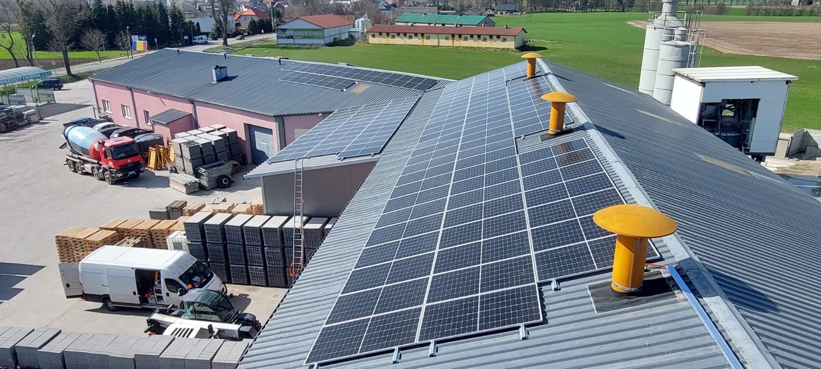 Instalacja fotowoltaiczna na dachu budynku przemysłowego z trzema żółtymi wywietrznikami, widok z góry na zaplecze z betoniarką i stertami kostki brukowej.