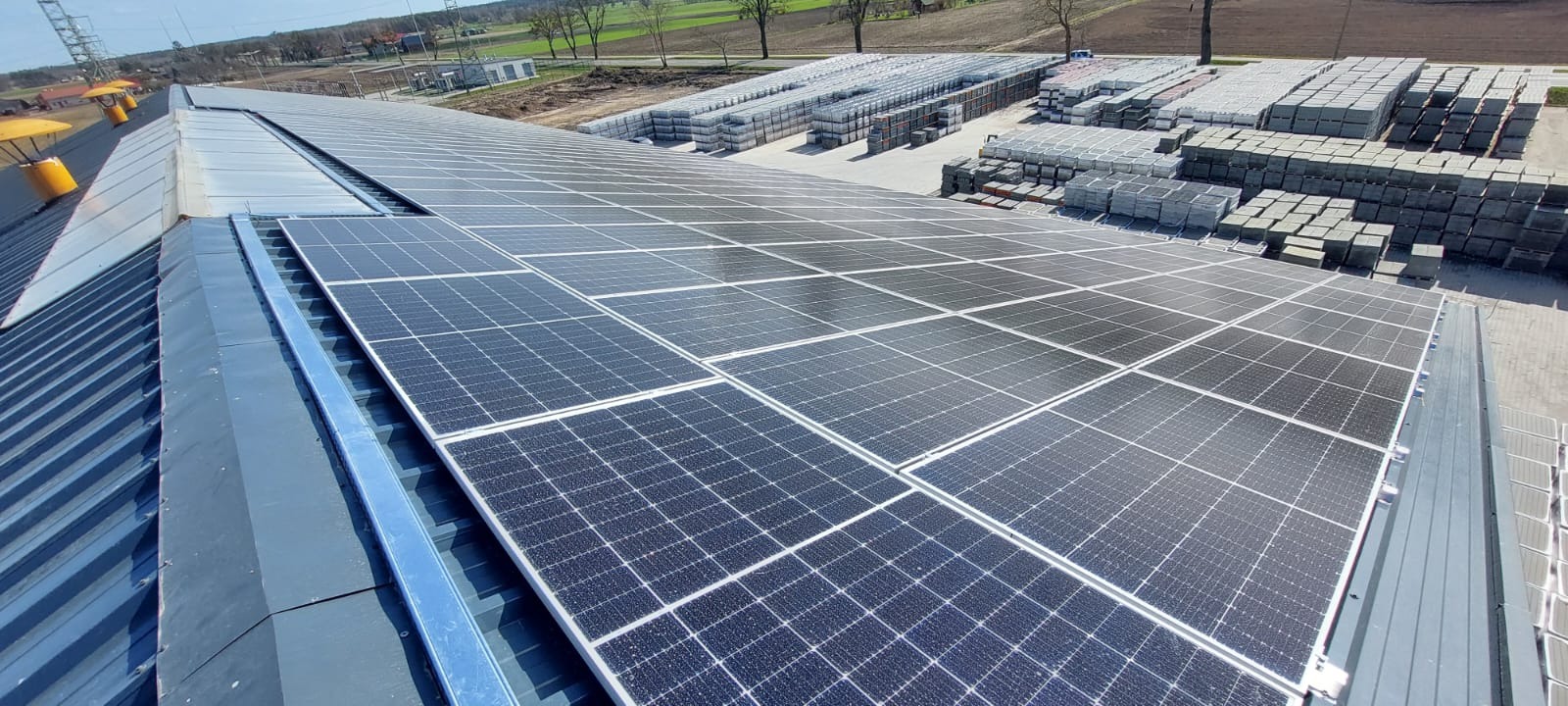 Rząd paneli słonecznych na dachu budynku przemysłowego, w tle składowane materiały budowlane.