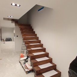 RomanBud - Drewniane schody dywanowe w trakcie montażu, zabezpieczone kartonem, widoczna drabina i materiały budowlane na palecie, surowe ściany i sufit z oświetleniem punktowym.