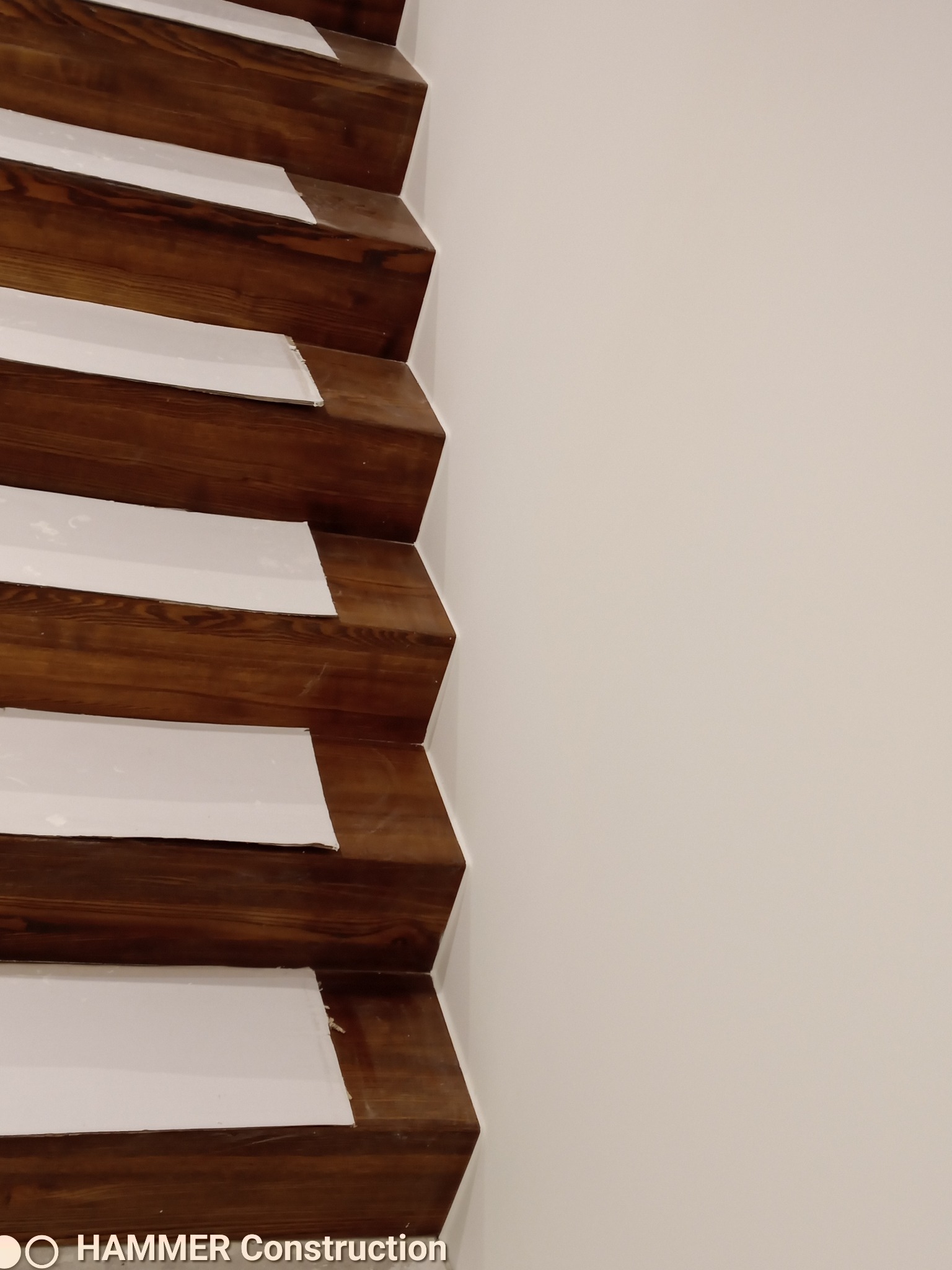 Drewniane schody z białymi podstopnicami w trakcie zabezpieczania przed pracami wykończeniowymi, widoczne nałożone taśmy malarskie, ściana w tle w kolorze złamanej bieli.