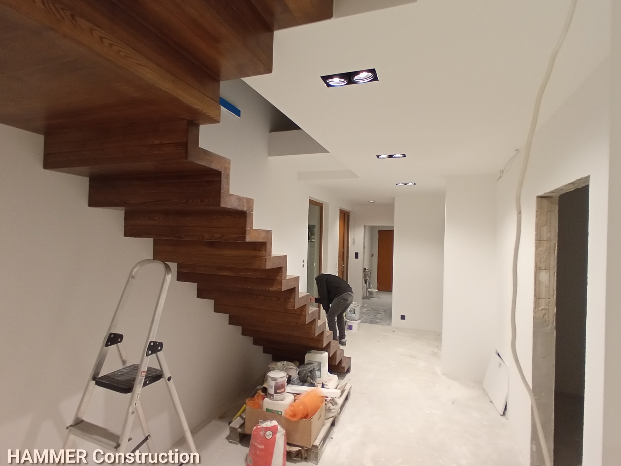 Montaż drewnianych schodów w nowoczesnym wnętrzu; widoczne materiały budowlane, drabina i pracownik wykonujący prace wykończeniowe.