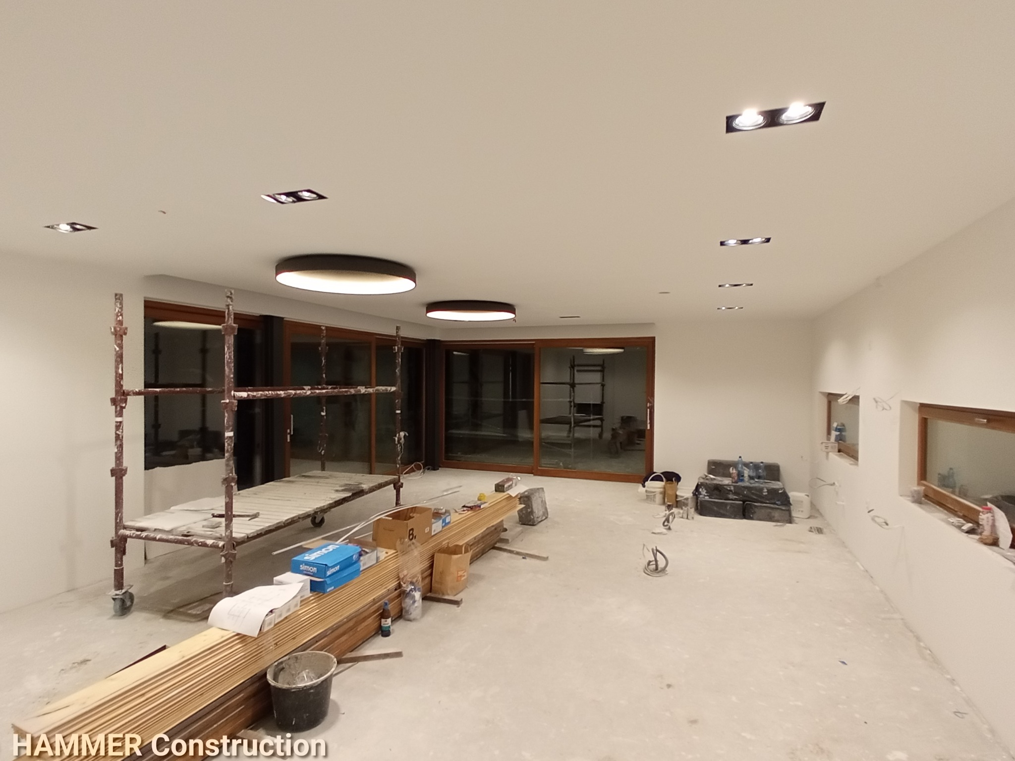 Wnętrze w trakcie prac wykończeniowych: widoczne rusztowanie, ułożone płyty kartonowo-gipsowe, narzędzia i materiały budowlane, duże okna przesuwne.