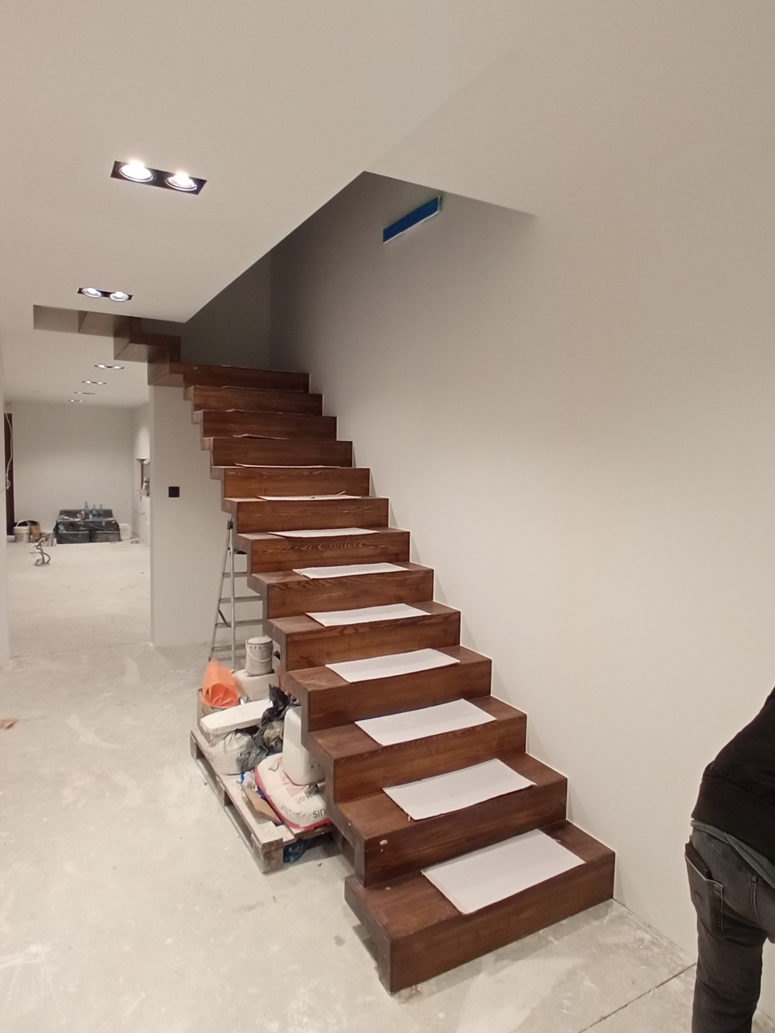 Drewniane schody dywanowe w trakcie montażu, zabezpieczone kartonem, widoczna drabina i materiały budowlane na palecie, surowe ściany i sufit z oświetleniem punktowym.