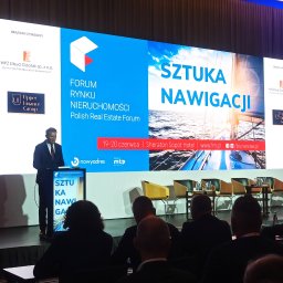 BSCAV SP. z o.o. - Prelegent w garniturze na scenie podczas Forum Rynku Nieruchomości, prezentacja ze zdjęciem żaglówki na ekranie, widownia w tle.