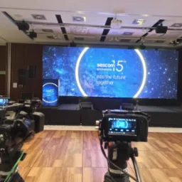 Profesjonalne kamery w studio filmowym ustawione przed sceną z dużym ekranem LED wyświetlającym logo firmy i hasło '15th anniversary into the future together'. Widoczne dodatkowe głośniki...