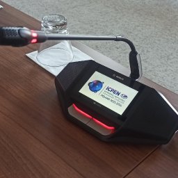 BSCAV SP. z o.o. - Czarny mikrofon konferencyjny Bosch z ekranem wyświetlającym logo ICPEN na drewnianym stole, obok szklanka z wodą.