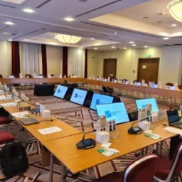 Sala konferencyjna w Gdyni, przygotowana do wydarzenia z monitorami wyświetlającymi logo, ustawionymi na stołach w kształcie podkowy. Widoczni uczestnicy i sprzęt konferencyjny.
