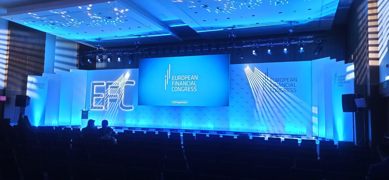 Scena konferencyjna z niebieskim oświetleniem, ekranem wyświetlającym logo i nazwę European Financial Congress oraz rzędami pustych krzeseł przed sceną. Widoczne reflektory i elementy dekoracyjne.