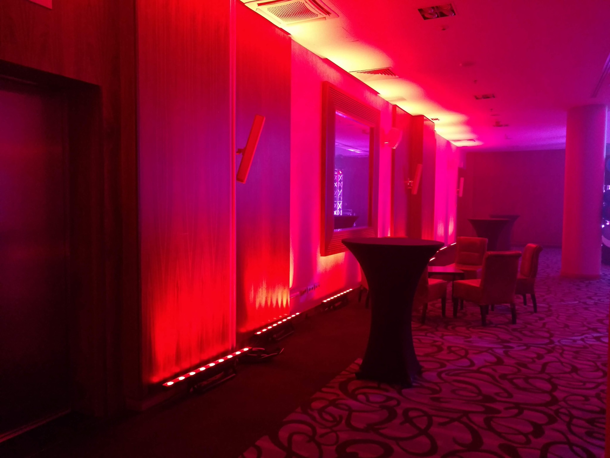 Korytarz oświetlony intensywnym czerwonym i różowym światłem, z czarnymi stolikami koktajlowymi i czerwonymi krzesłami, lustra na ścianach, dywan w abstrakcyjne wzory.