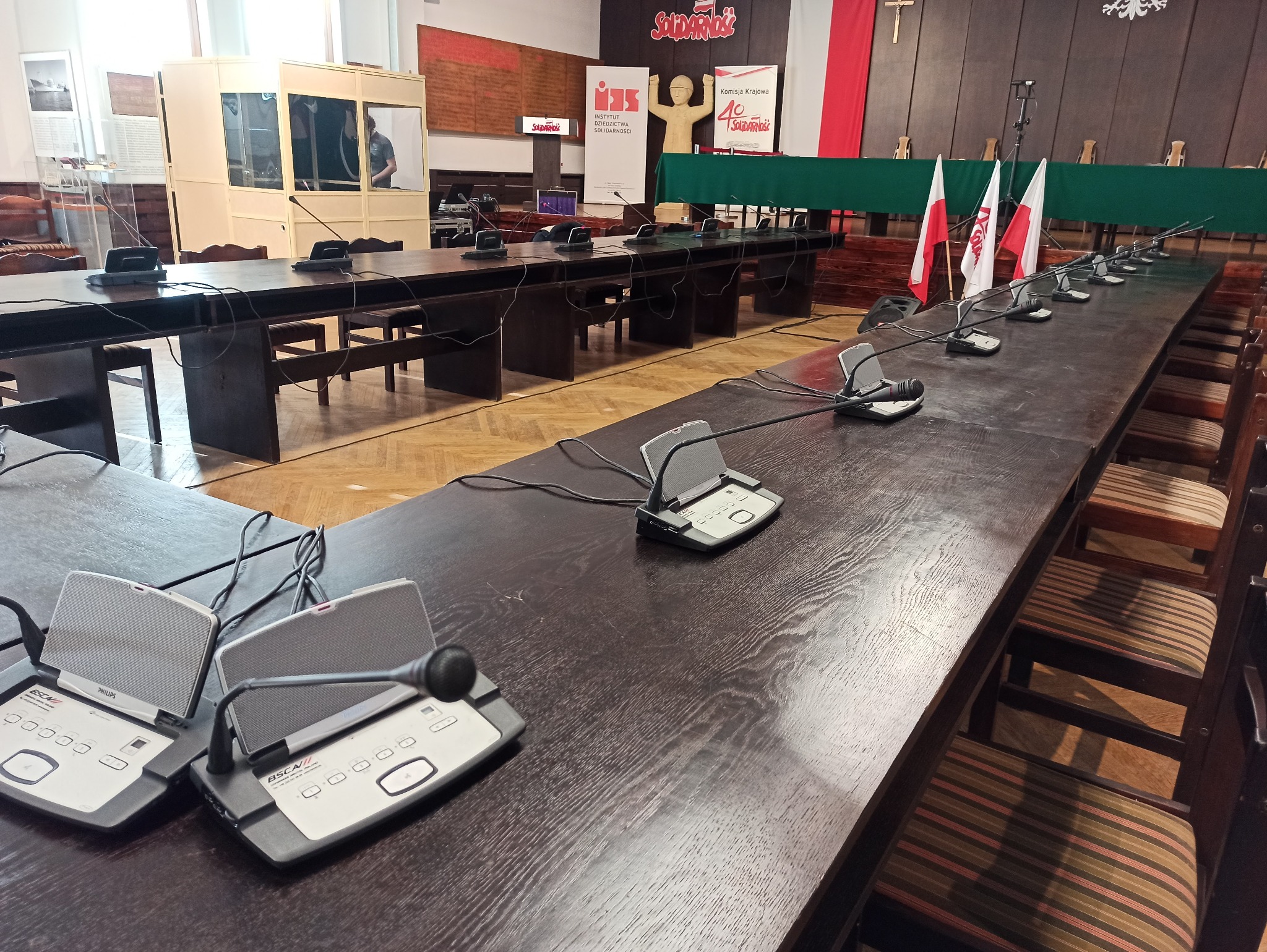 Sala konferencyjna z długim stołem, mikrofonami konferencyjnymi Philips BSCA/II, krzesłami i flagami Polski w tle. Na ścianie plakaty związane z Solidarnością.