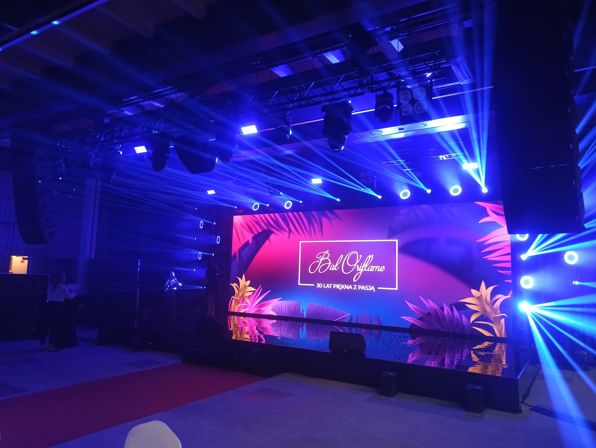 Scena z ekranem wyświetlającym logo 'Bal Oriflame' i hasło '30 lat piękna z pasją', oświetlona niebieskimi reflektorami, z widocznym nagłośnieniem i dekoracjami roślinnymi.