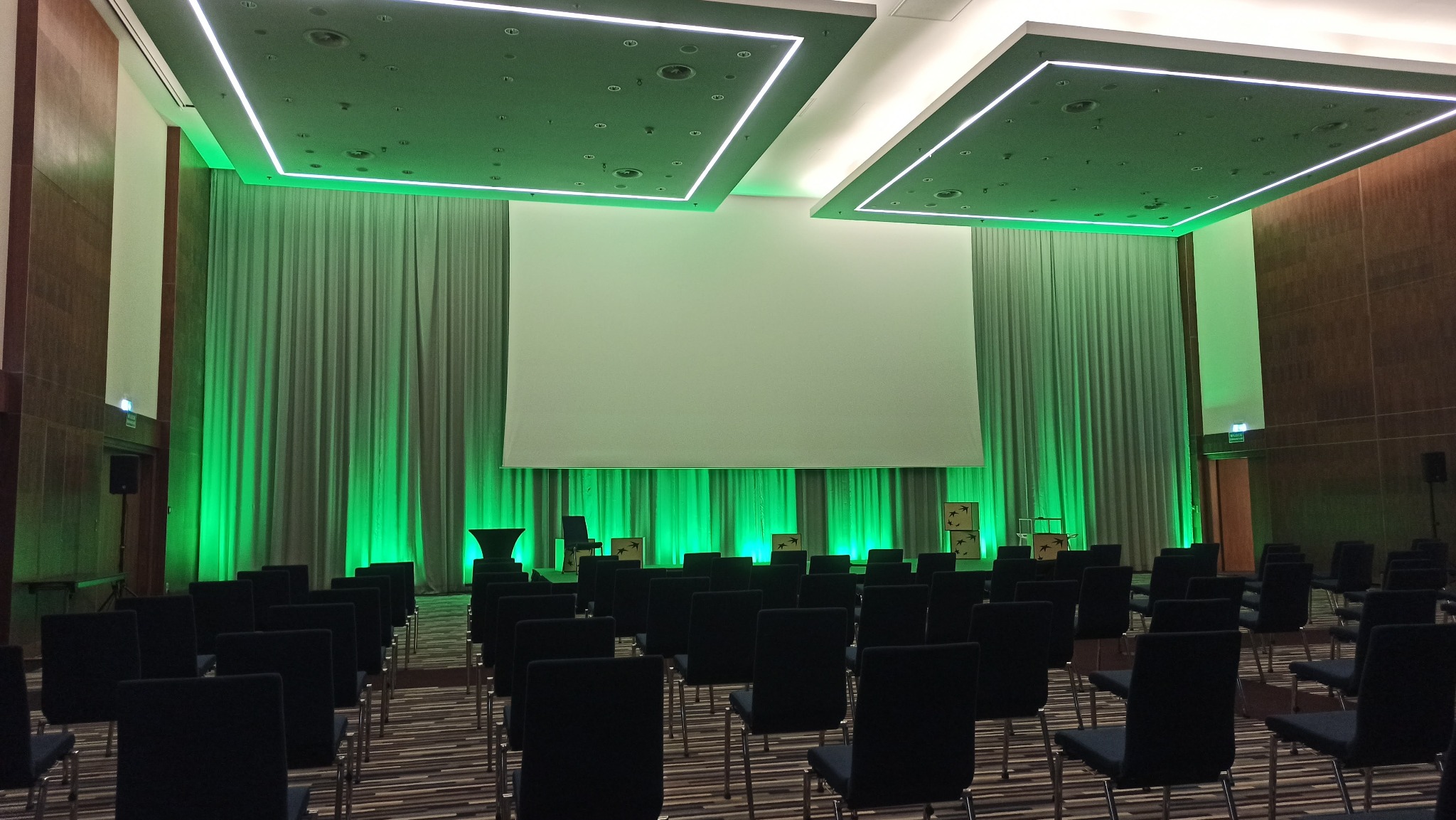 Sala konferencyjna z rzędami krzeseł, ekranem projekcyjnym i zielonym oświetleniem, widoczne kolumny głośnikowe i dekoracje na scenie.