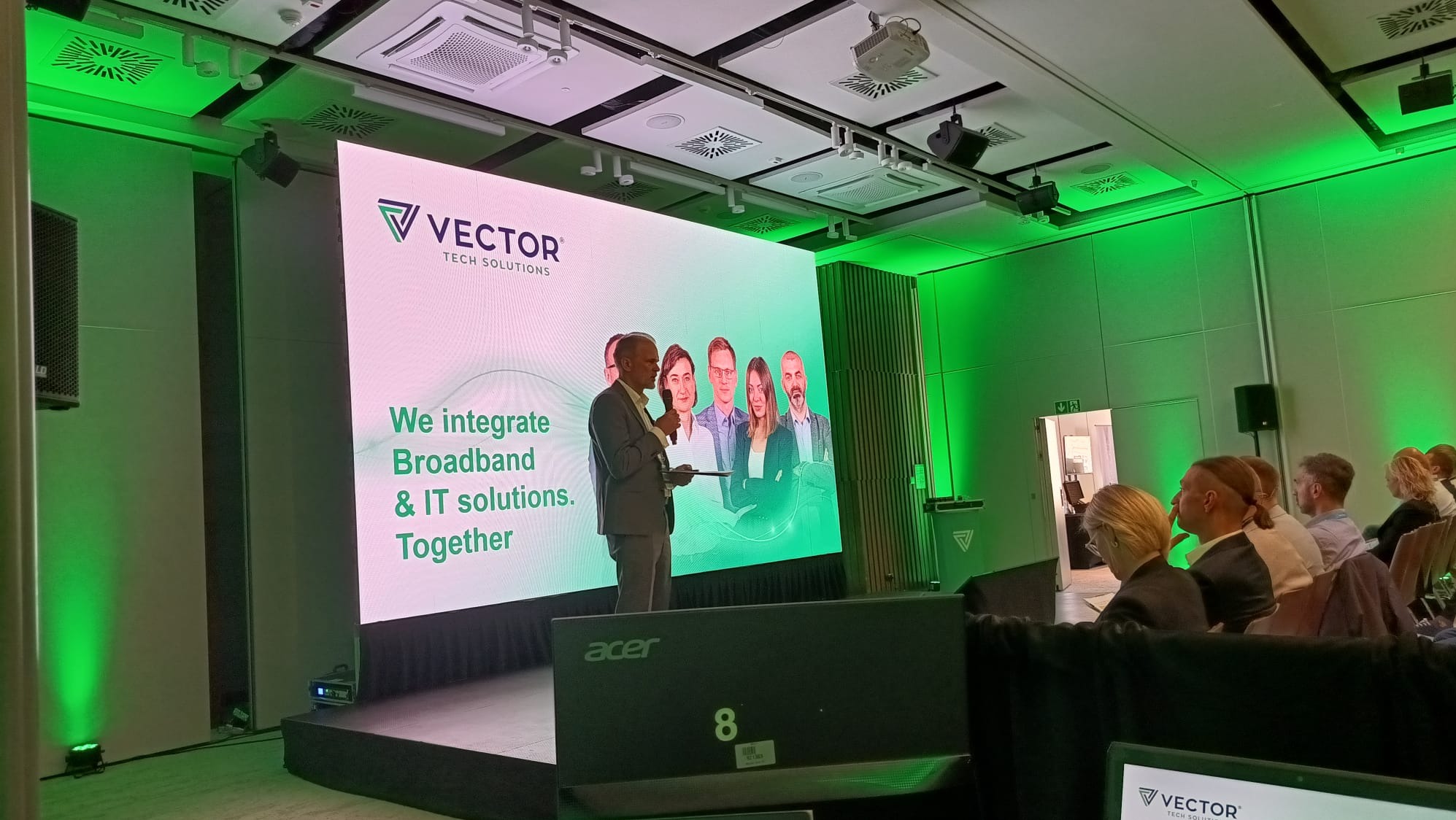 Prelegent w szarym garniturze przemawia na scenie z mikrofonem podczas konferencji, na ekranie LED wyświetlane logo firmy Vector Tech Solutions i zdjęcia zespołu, zielone oświetlenie sceniczne...