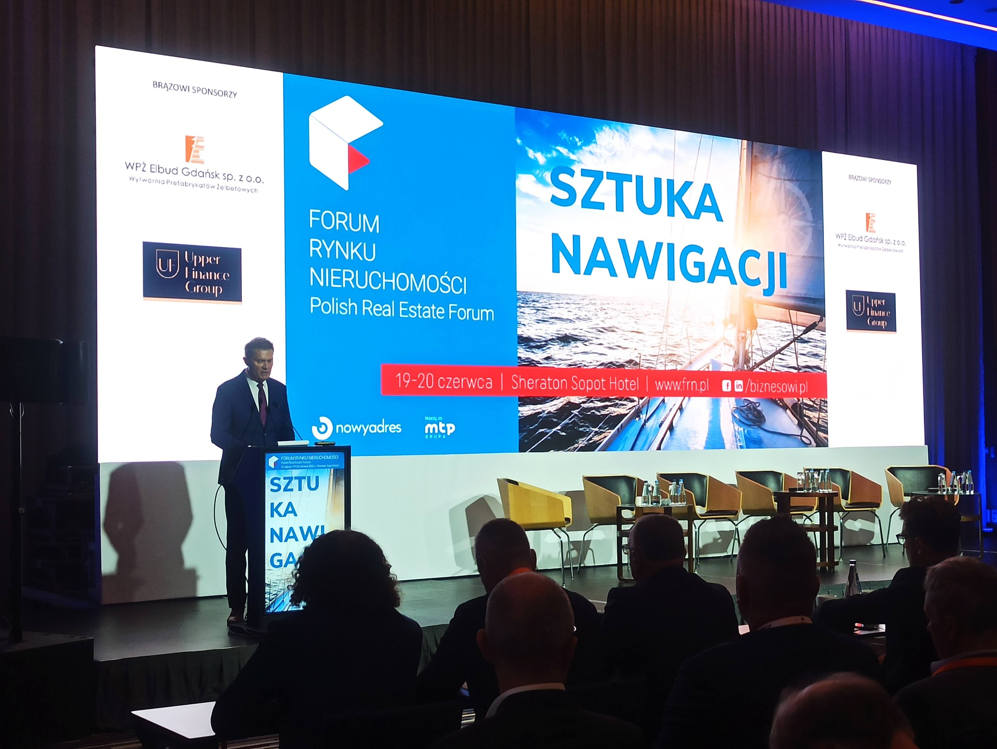 Prelegent w garniturze na scenie podczas Forum Rynku Nieruchomości, prezentacja ze zdjęciem żaglówki na ekranie, widownia w tle.